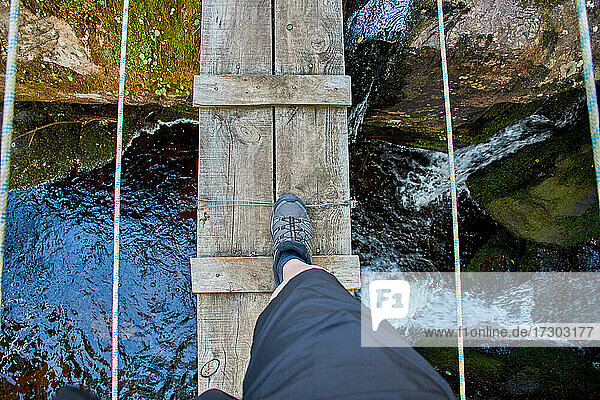 Das Bein eines Sportlers  der eine Holzbrücke mit Seilen über ein natürliches Felsbecken und einen Wasserfall überquert.