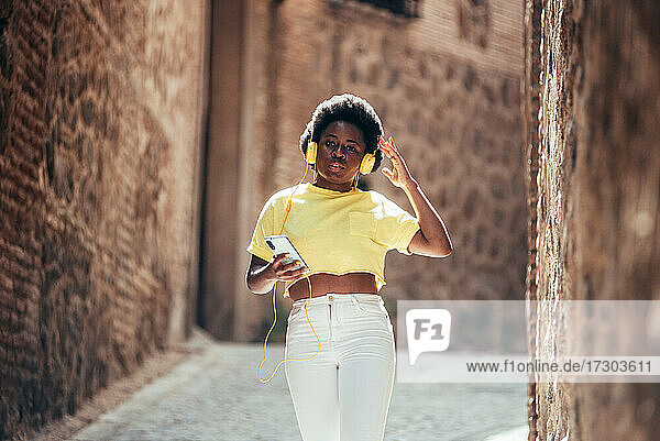 Porträt eines schwarzen Mädchens mit Afrofrisur  das mit Kopfhörern und ihrem Handy Musik hört  während es eine Straße in der Altstadt entlanggeht.