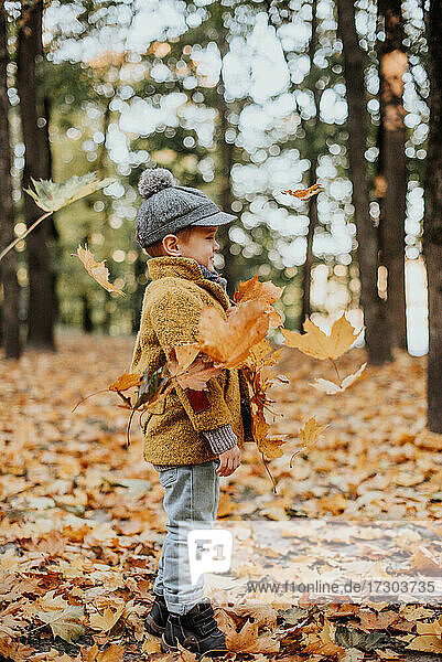 Stylish boy having fun in autumn city park. Happy kid playing woth fallen leaves. Kids fashion. Boy wearing trendy yellow coat  cap and scarf. Smiling young boy outdoors. Kid jumping and runn