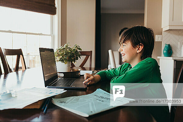 Happy boy doing on school work online on computer at kitchen table.