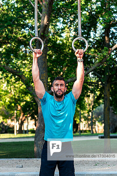 Dark-haired athlete with beard holding on to gymnastic rings.