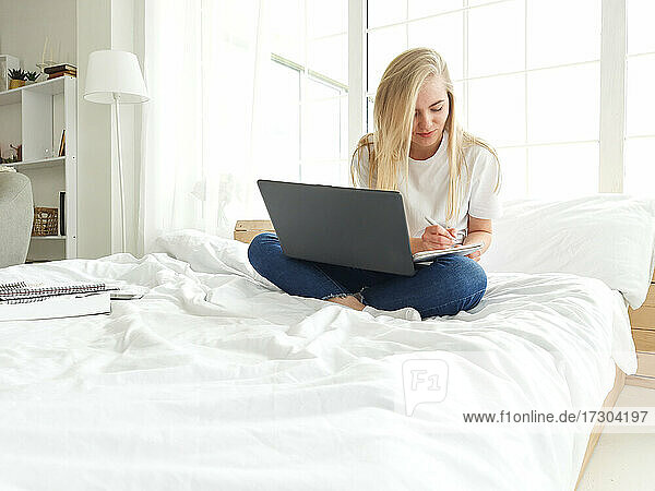 blonde junge Frau macht sich Notizen mit Stift mit Latop auf ihren Nüssen im Bett