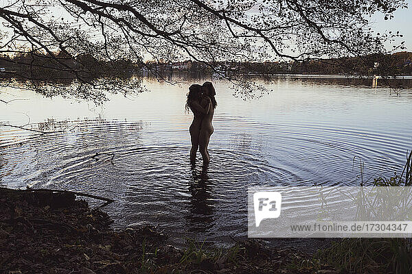 lesbische queer Paar umarmen nackt in plätschernden See Wasser unter Baum