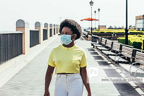 Porträt eines schwarzen Mädchens mit Gesichtsmaske  Afrohaar und Ohrringen  das durch einen städtischen Raum geht.