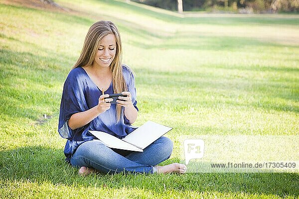 Lächelnde junge Frau mit Buch und Handy im Freien im Park