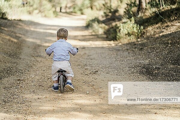 Back of cute toddler riding bicycle on dirt road in forest