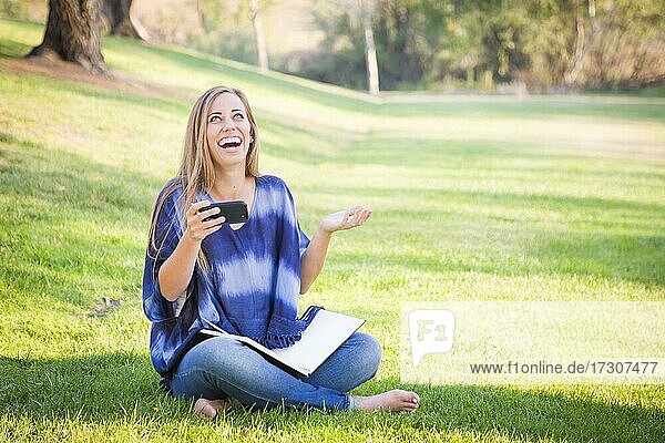 Lachende junge Frau mit Buch und Mobiltelefon im Freien im Park
