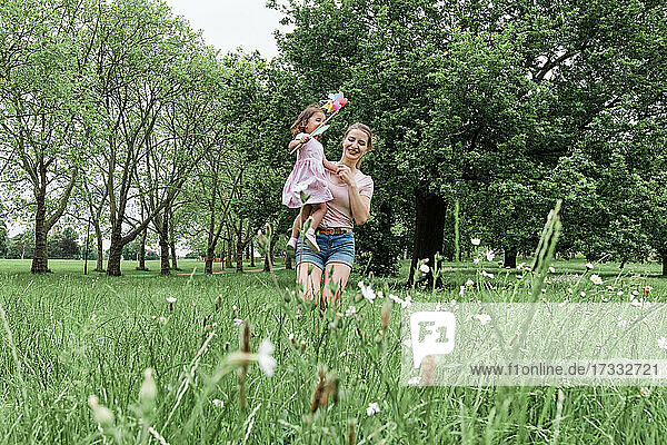 Glückliche Mutter  die ihre Tochter mit einem Windrad trägt  während sie im Park auf dem Rasen spazieren geht