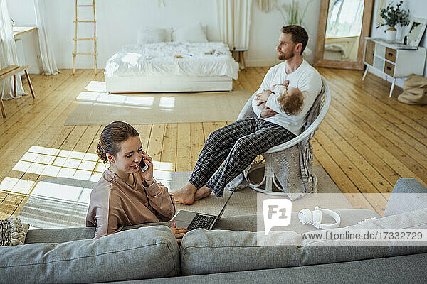 Geschäftsfrau mit Laptop  die mit einem Mobiltelefon spricht  während der Mann seine Tochter auf einem Stuhl zu Hause trägt