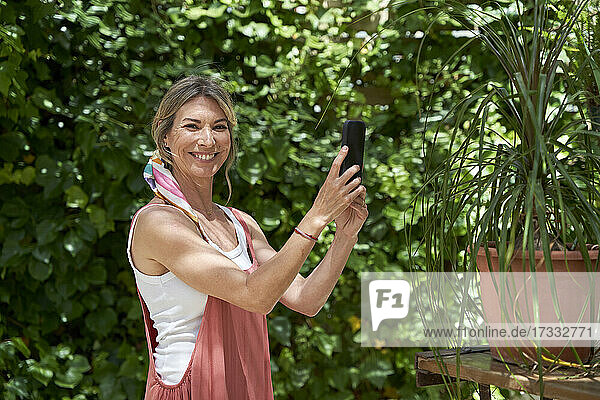 Lächelnde Frau  die mit ihrem Smartphone Pflanzen fotografiert  während sie im Hinterhof steht