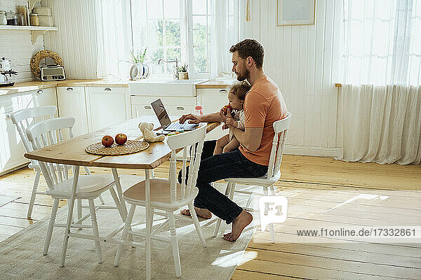 Businessman feeding daughter while working on laptop at home