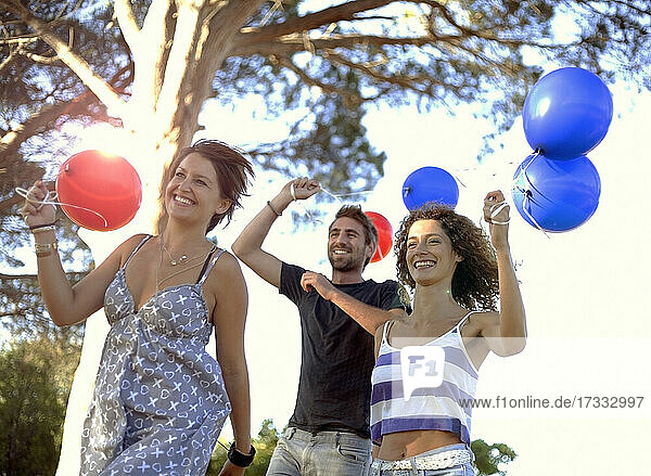 Unbeschwerte männliche und weibliche Freunde laufen mit roten und blauen Luftballons in der Hand