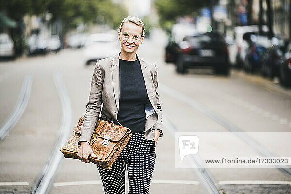 Lächelnde Geschäftsfrau mit Hand in der Tasche  die eine Tasche hält  während sie auf der Straße geht