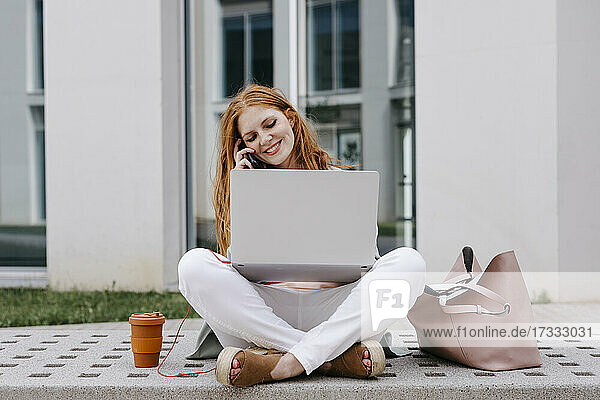 Businesswoman sitting cross-legged on bench talking on smart phone while using laptop
