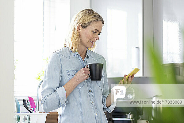 Frau mit Tasse  die in der Küche zu Hause ein Mobiltelefon benutzt