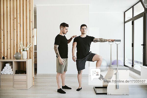 Man exercising at stability chair while male instructor supporting at pilates studio