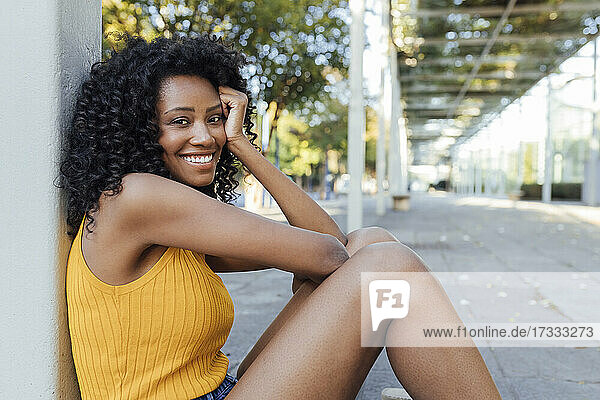 Lächelnde junge Frau mit lockigem Haar an einer Säule sitzend