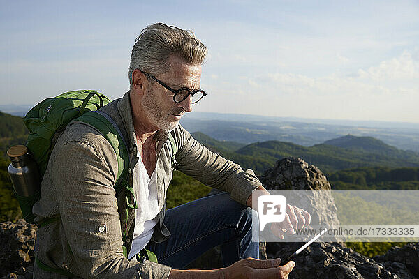 Männlicher Wanderer  der auf einem Berg sitzend ein digitales Tablet benutzt