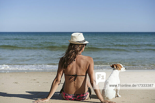 Frau im Bikini mit Hut sitzt mit Hund am Strand