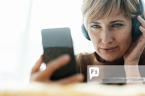 Ältere Geschäftsfrau hört Musik über Kopfhörer  während sie ihr Smartphone im Büro benutzt