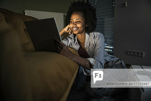 Lächelnde Frau  die ein digitales Tablet benutzt  während sie sich zu Hause auf dem Sofa ausruht