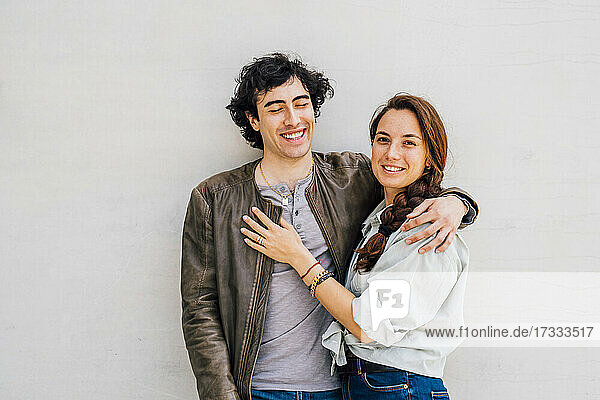 Lächelndes Paar mit umschlungenen Armen vor einer Wand stehend