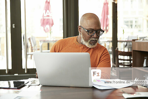 Glatzköpfiger Geschäftsmann  der in einem Restaurant an einem Laptop arbeitet und Dokumente betrachtet