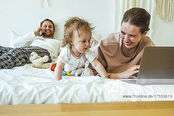 Lächelnde Frau mit Laptop  die ihre Tochter anschaut  während der Mann im Schlafzimmer mit dem Handy telefoniert