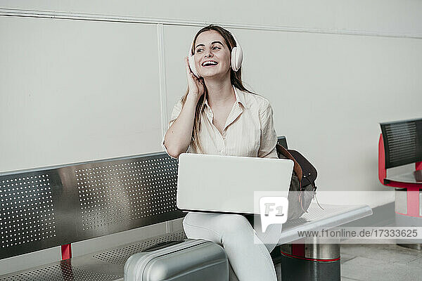 Fröhliche junge Frau  die mit einem Laptop am Bahnhof sitzt und Musik über Kopfhörer hört