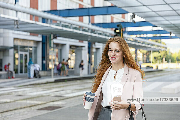 Lächelnde Geschäftsfrau mit Smartphone  die am Bahnhof wegschaut