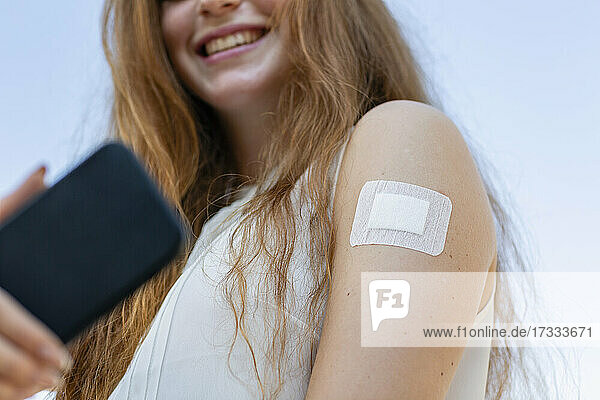 Lächelnde Geschäftsfrau mit Impfstoff am Arm macht ein Selfie mit ihrem Smartphone während der COVID-19