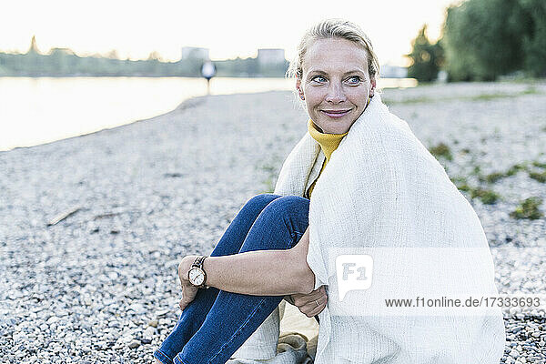 Lächelnde Frau in Decke eingewickelt auf einem Kieselstein sitzend