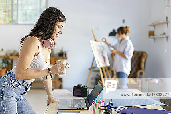 Weibliche Fotografin hält einen Becher  der auf einem Schreibtisch steht  während ein Künstler im Hintergrund malt