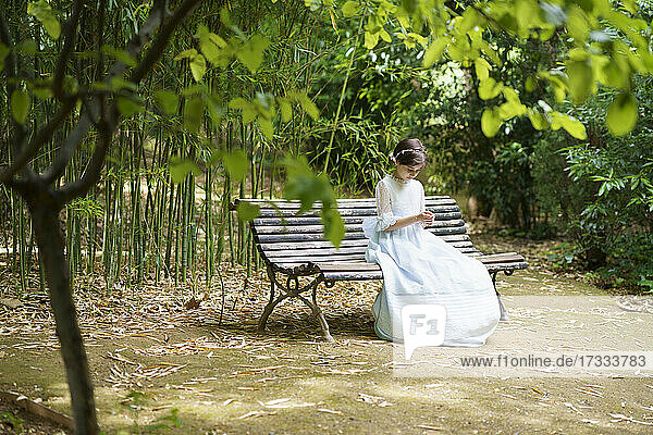 Girl in white communion dress sitting on bench in park