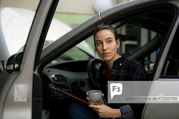 Mechanikerin mit Kaffeetasse  die wegschaut  während sie im Auto sitzt