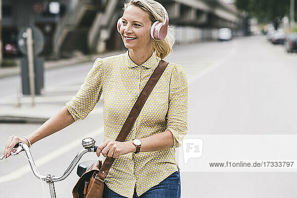 Blonde Geschäftsfrau mit Kopfhörern lächelt  während sie auf der Straße Fahrrad fährt