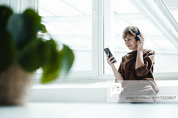 Geschäftsfrau mit Kopfhörern und Smartphone am Fenster sitzend