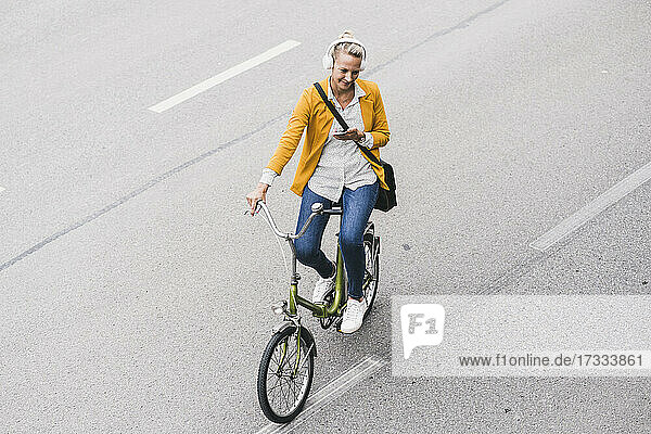Ältere Geschäftsfrau mit Kopfhörern und Mobiltelefon fährt Fahrrad auf der Straße