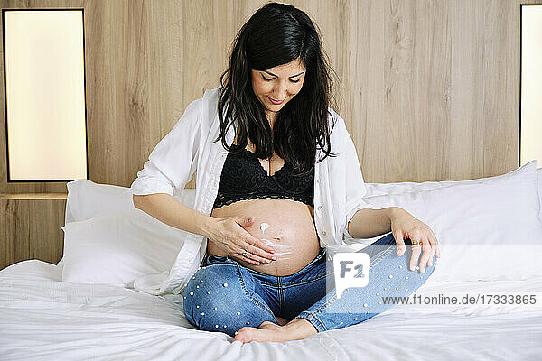Schwangere Frau trägt Feuchtigkeitscreme auf den Bauch auf  während sie auf dem Bett im Schlafzimmer sitzt