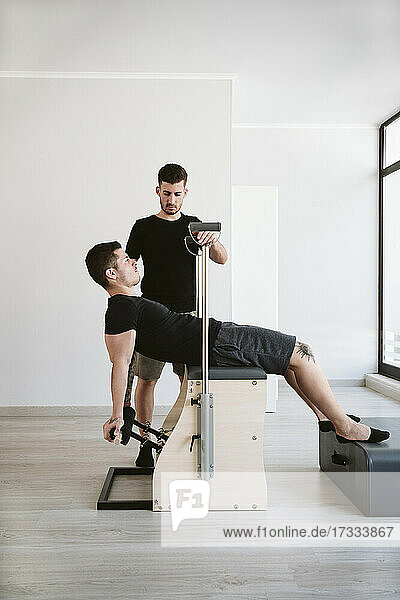 Männlicher Fitnesslehrer  der einen Mann betrachtet  der sich auf einem Stabilitätsstuhl im Pilates-Studio dehnt
