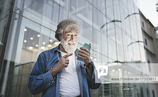 Happy mature man pointing at smart phone in front of glass building