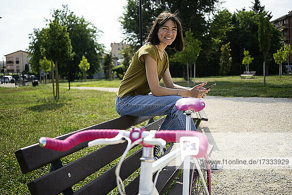 Lächelnde Frau  die auf einer Bank in der Nähe eines Fahrrads auf dem Fußweg sitzt