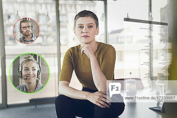 Businesswoman with hand on chin sitting on desk with male and female colleague icons in corner