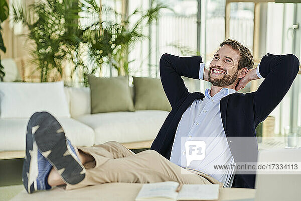 Fröhlicher männlicher Freiberufler  der mit hochgelegten Füßen auf dem Tisch im Heimbüro sitzt und wegschaut