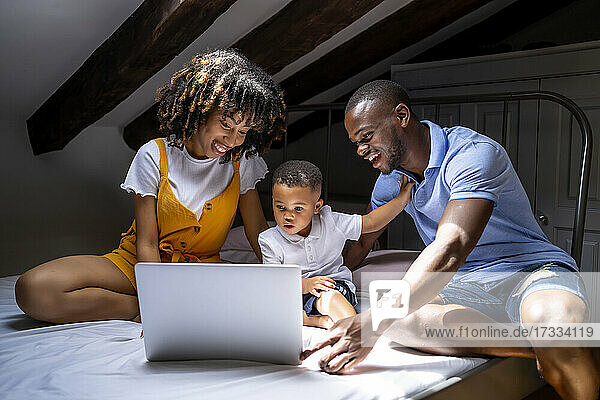 Glückliche Familie mit Sohn auf dem Bett sitzend und Laptop benutzend auf dem Dachboden zu Hause
