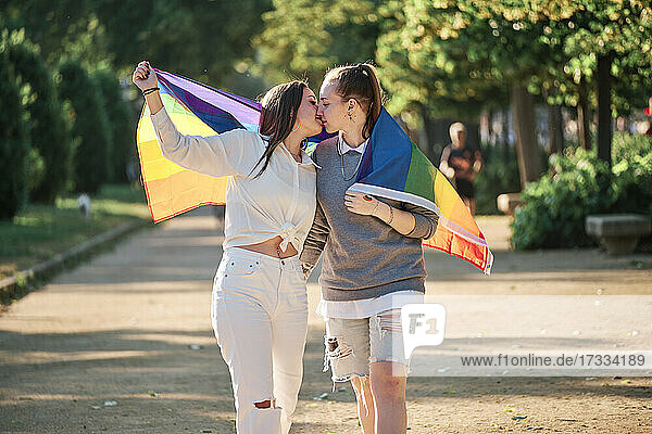 Paar mit Regenbogenflagge küsst sich beim Spaziergang im Park