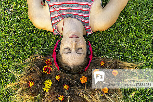 Young woman with eyes closed listening music through headphones with flowers in hair lying on grass
