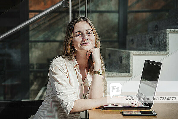 Lächelnde junge Geschäftsfrau sitzt mit der Hand am Kinn am Tisch in einem Café