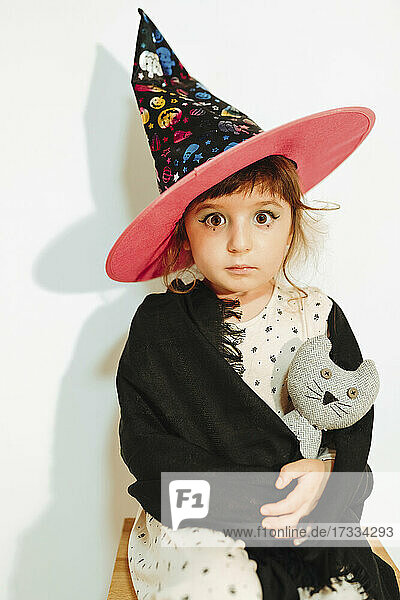 Girl dressed as Halloween witch holding stuffed toy by wall