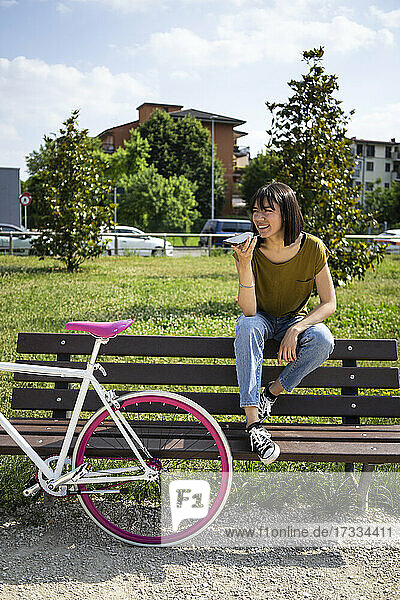 Junge Frau mit Fahrrad  die eine Sprachnachricht über ihr Smartphone sendet  während sie auf einer Bank im Park sitzt
